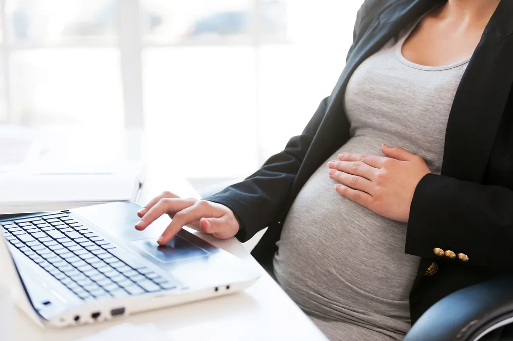 A pregnant woman resting her hand on her stomach while working on her laptop.