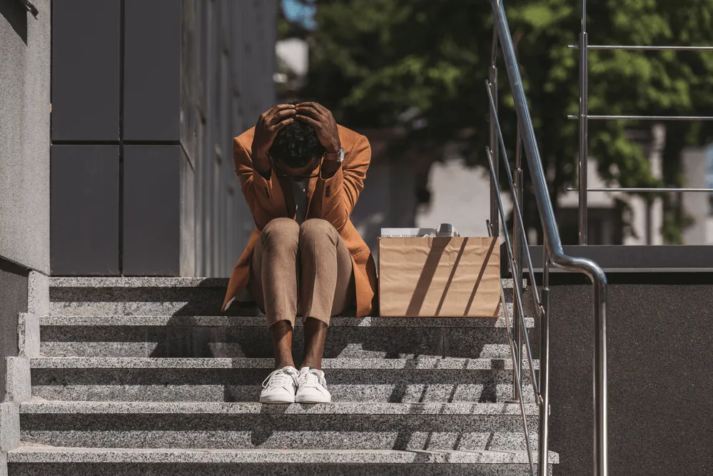 A person sits on steps looking down and clutching their head with a cardboard box of belongings next to them.