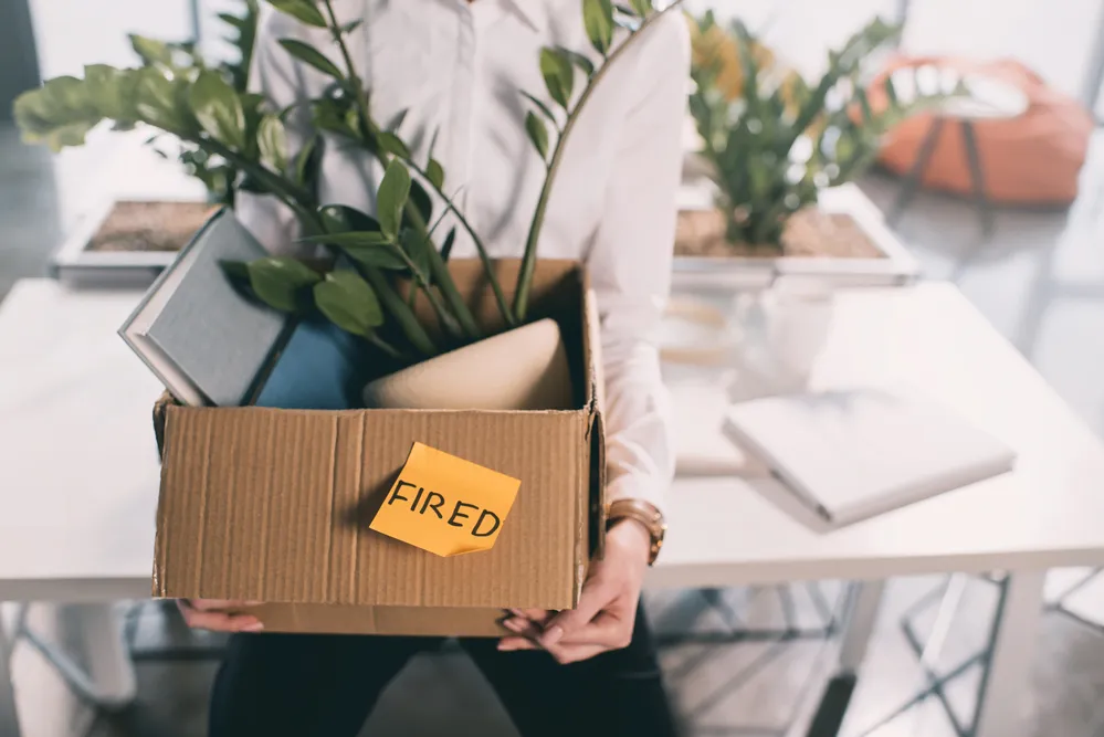A person holding a cardboard box containing a plant and books with a post it note on it that reads fired.