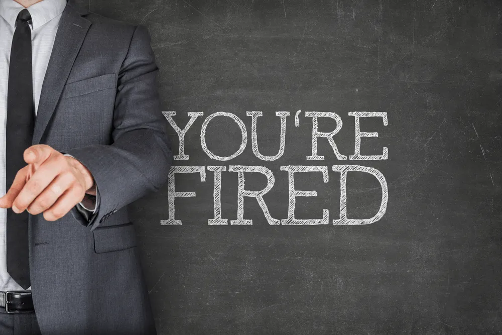 A man in a suit pointing towards the camera with a chalkboard behind him that says You're Fired.
