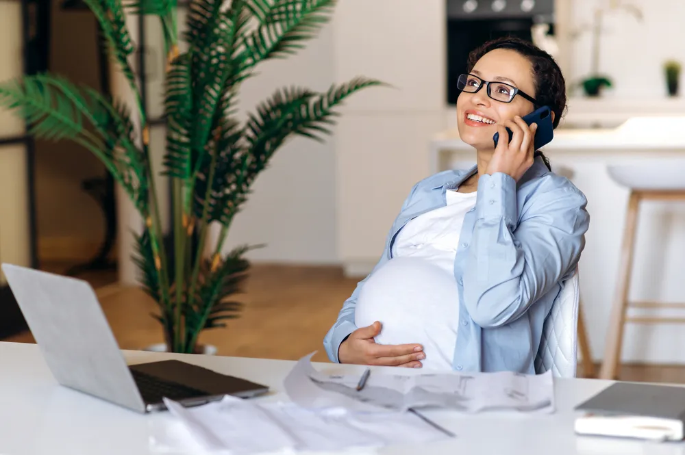 A pregnant woman sitting at her desk talking on a cell phone.